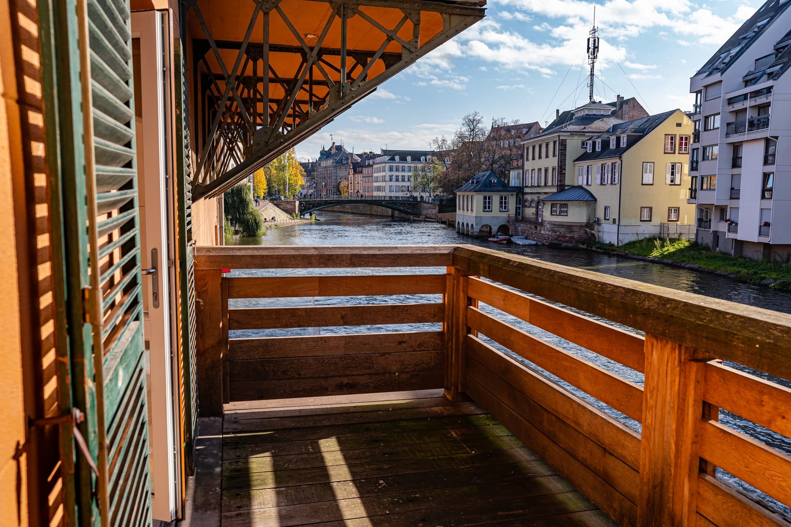Le balcon de bois du Moulin aux Épices, suspendu au-dessus de la rivière, volet ouvert, face aux quais de Strasbourg