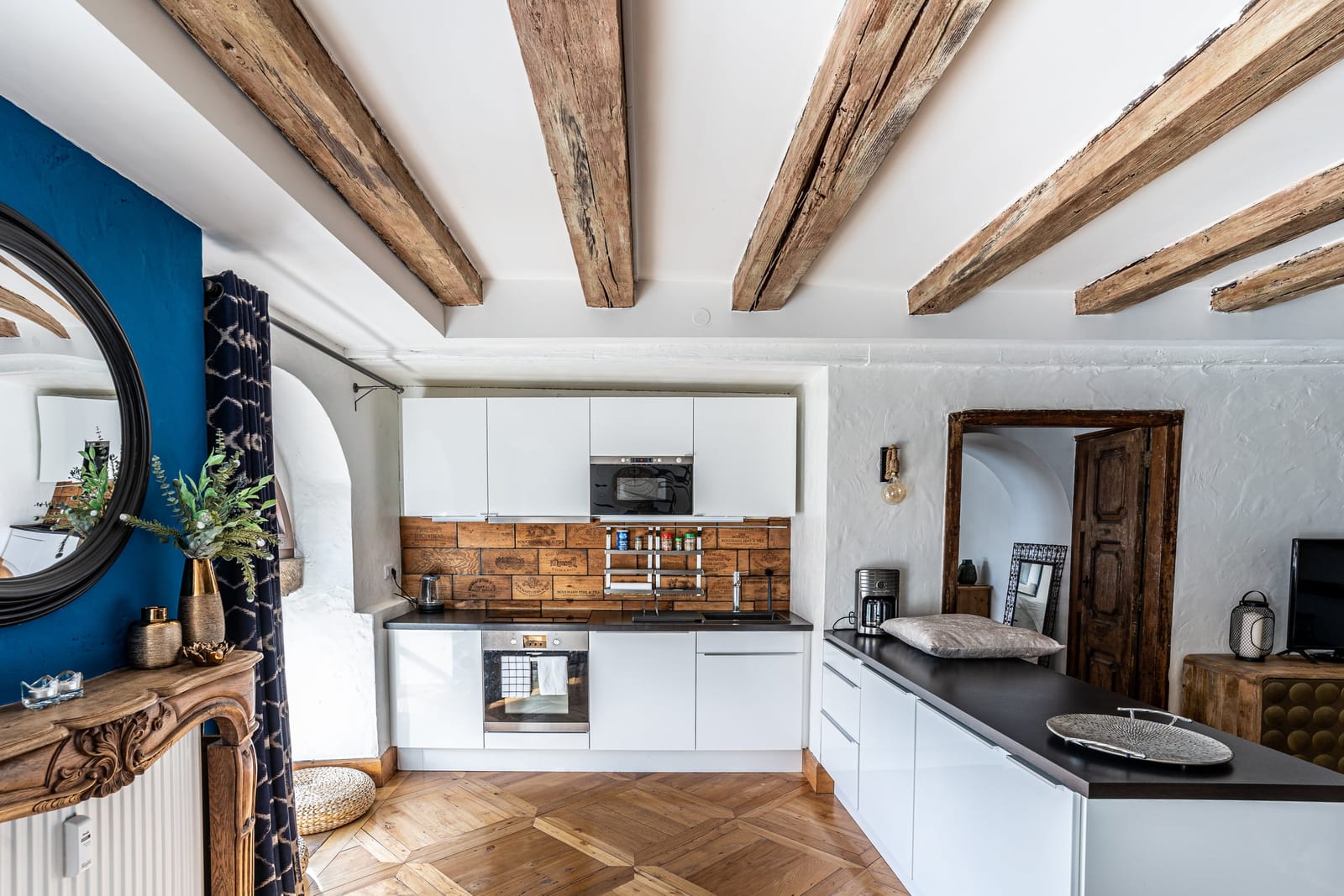La cuisine du Moulin aux Épices sous les poutres du moulin d'origine : mur bleu, miroir rond, cuisine blanche et parquet ancien