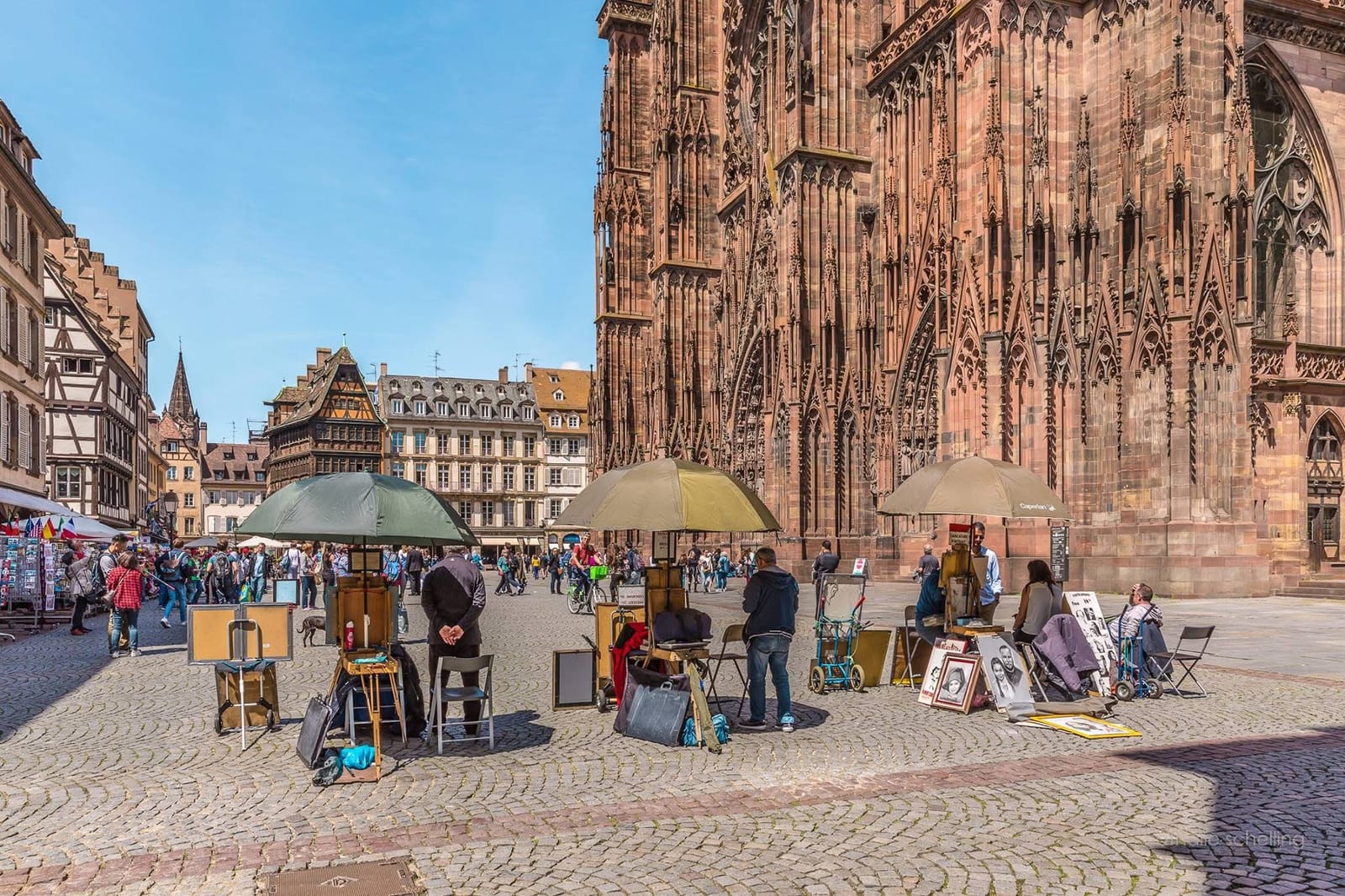 Le pied de la cathédrale en été : la place animée, les dessinateurs et leurs parasols devant le grès rose