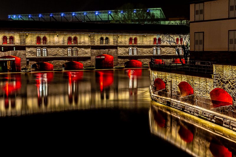 Le barrage Vauban illuminé la nuit, ses reflets rouges dans l'Ill