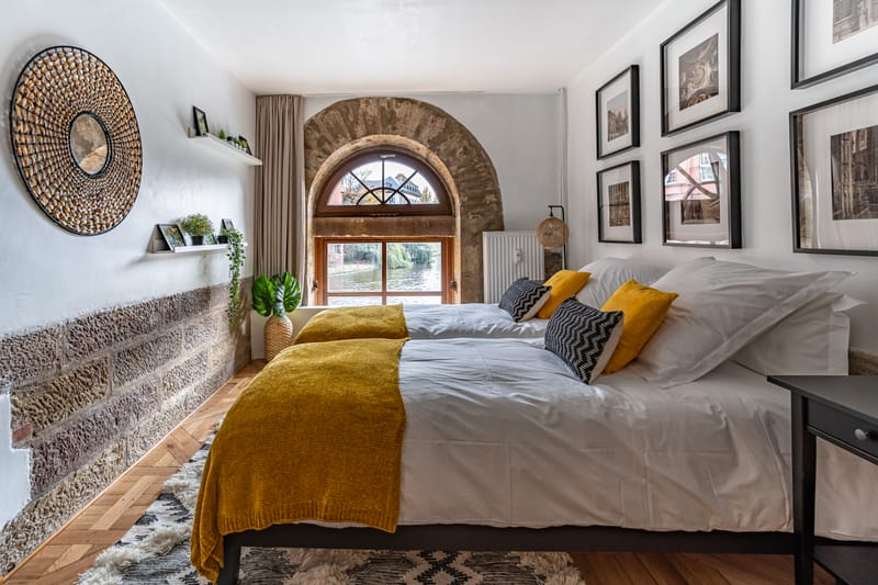 Une chambre sous les poutres : lit face au mur de pierre, fenêtre en plein cintre et cadres au mur