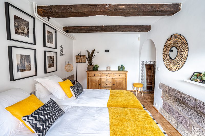 La chambre sous les poutres : mur de pierre, miroir en rotin et coussins jaunes