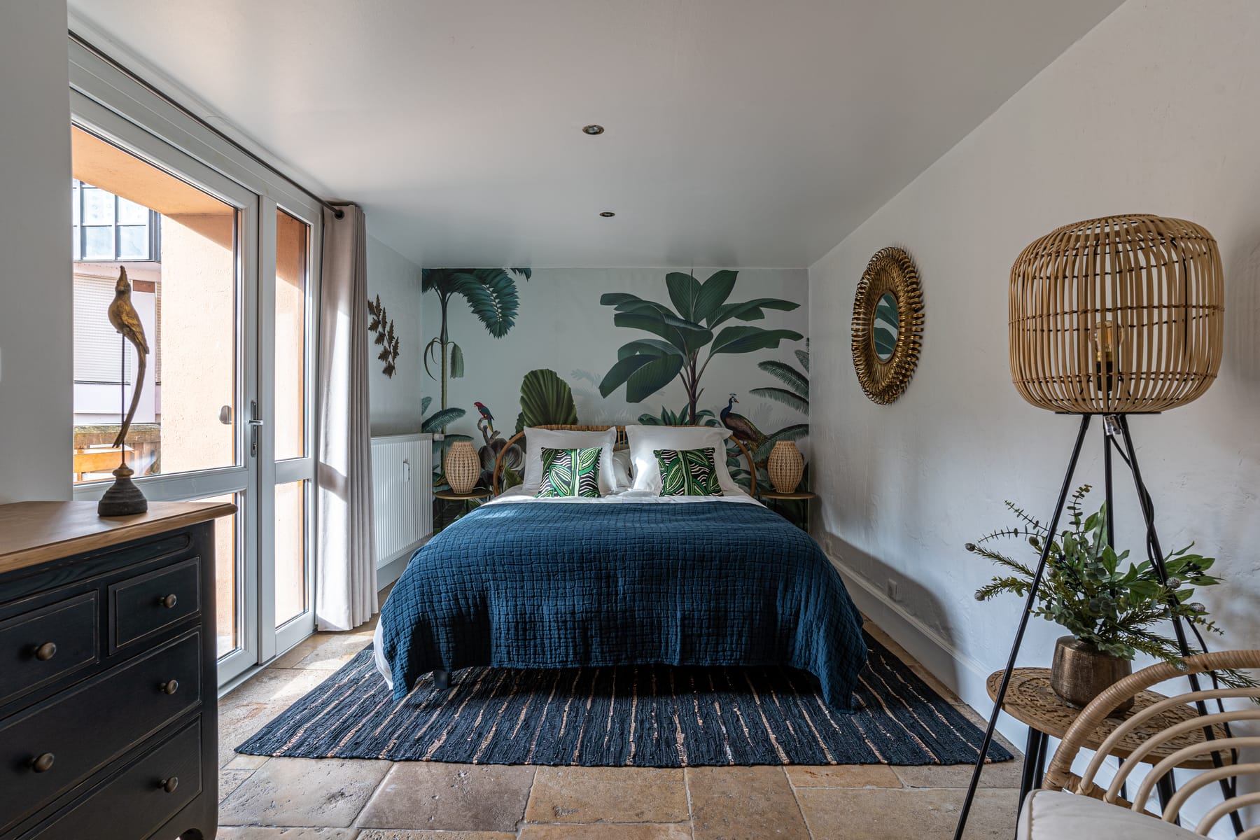 La seconde chambre du Moulin aux Épices : grand lit au couvre-lit bleu nuit, papier peint de feuillages, lampe en rotin et porte vitrée