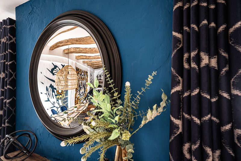 Le miroir rond sur le mur bleu, entre les rideaux à motifs, et le reflet des poutres