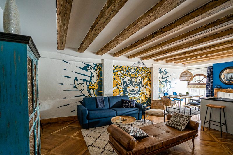 Le séjour vu de l'angle : buffet bleu, canapé, banquette de cuir et la fresque sous les poutres