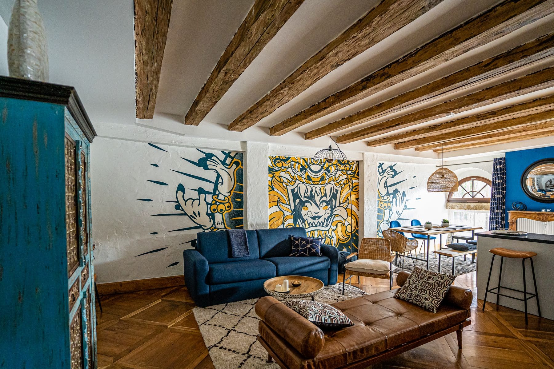 Le séjour du Moulin aux Épices sous les poutres : la fresque murale en bleu et or, le canapé bleu, et au fond les fenêtres en plein cintre