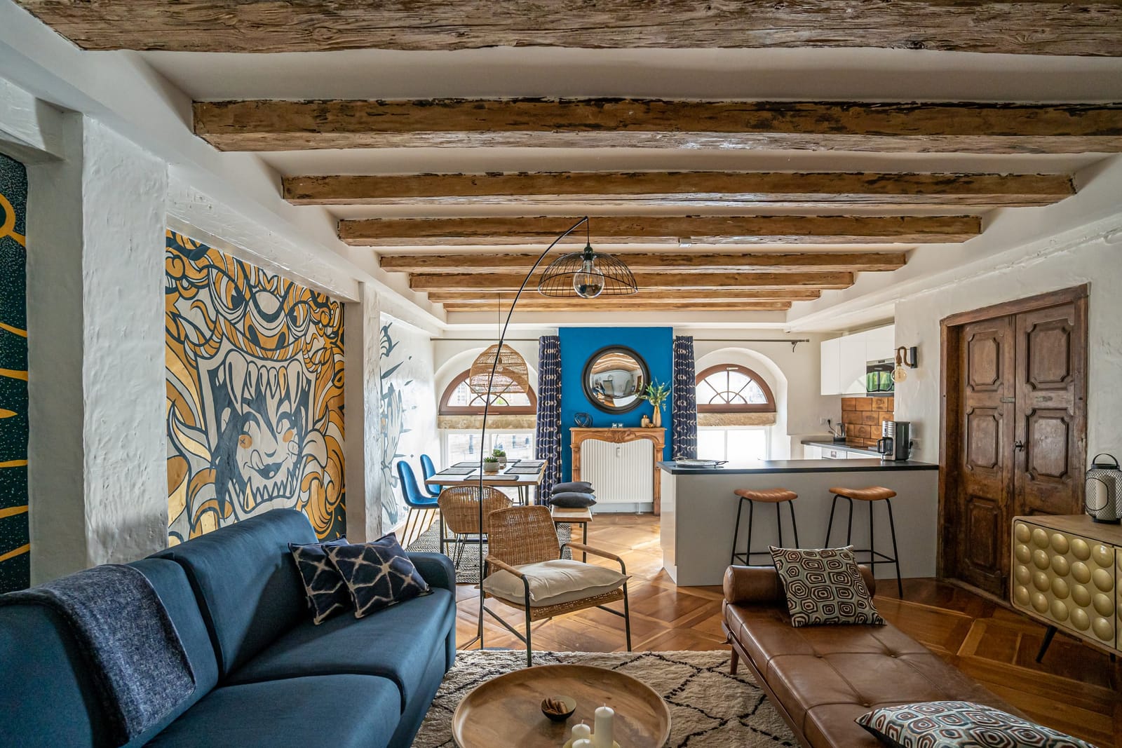 Le séjour du Moulin aux Épices sous les poutres : canapé bleu, fresque murale, et au fond les fenêtres en plein cintre et la cuisine